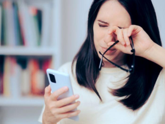 O imagine care arată o femeie de 30 de ani privindu-și telefonul în timp ce se freacă la ochi.