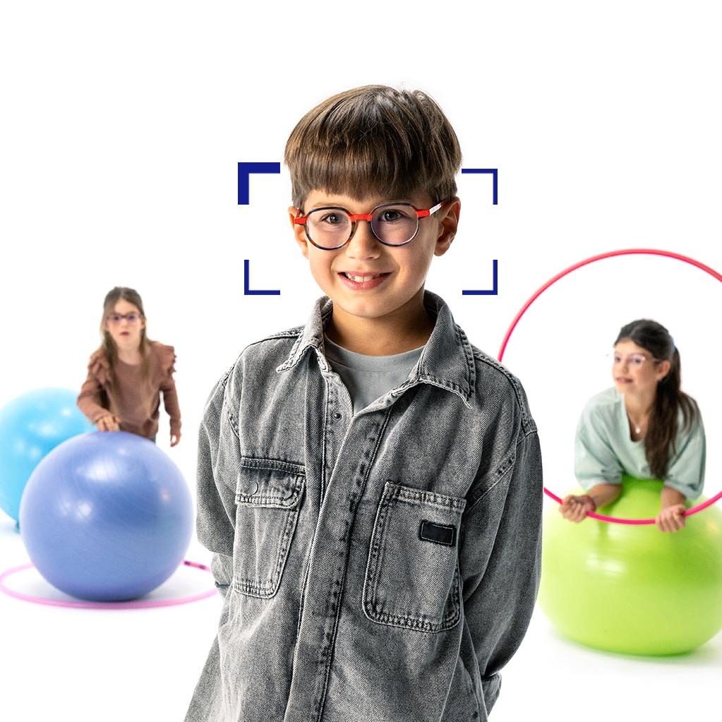 Băiat cu părul șaten cu ochelari rotunzi, purtând lentilele ZEISS MyoCare, stă în prim-plan și zâmbește la cameră. În fundal sunt două fete care poartă lentile ZEISS MyoCare care se joacă cu cercuri și mingi de gimnastică.