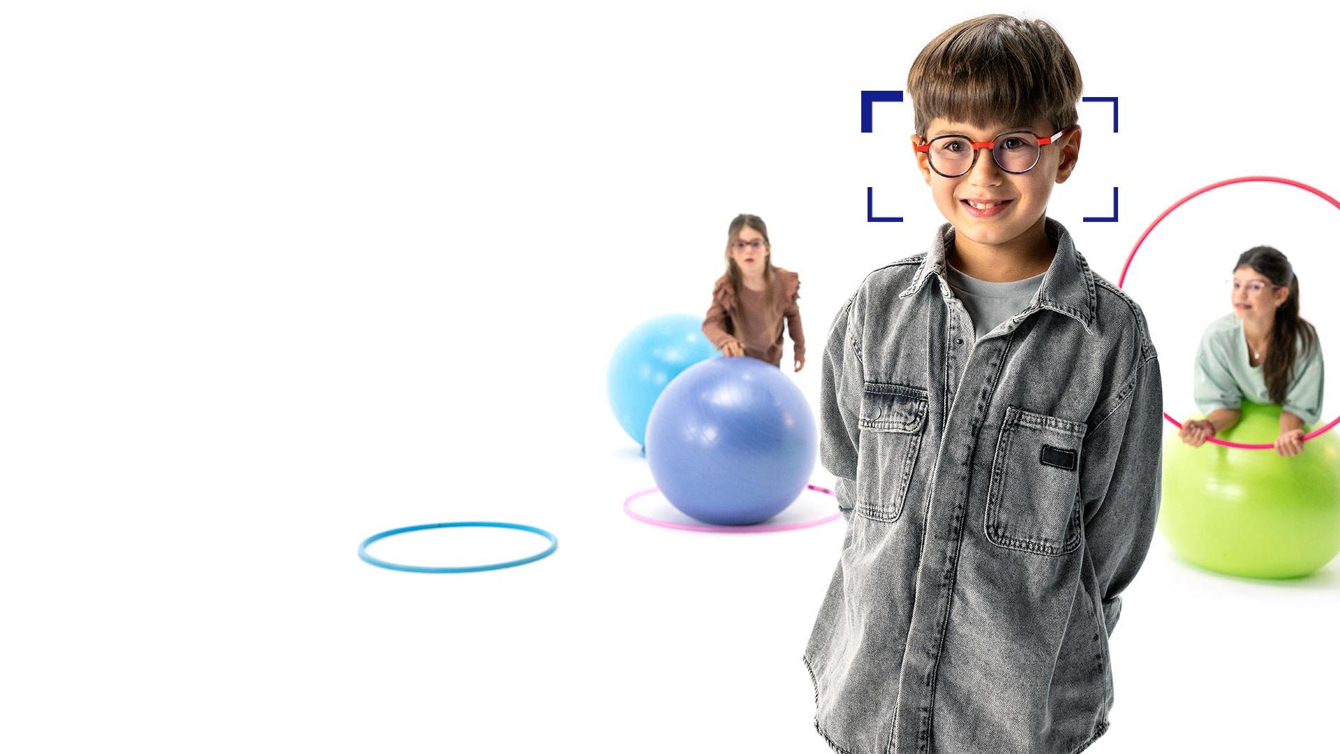 Băiat cu părul șaten cu ochelari rotunzi, purtând lentilele ZEISS MyoCare, stă în prim-plan și zâmbește la cameră. În fundal sunt două fete care poartă lentile ZEISS MyoCare care se joacă cu cercuri și mingi de gimnastică.