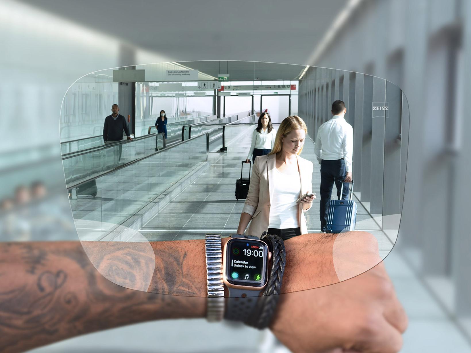 Persoană care se uită la ceasul de pe mână prin lentilele ZEISS SmartLife Digital. În fundal se vede o imagine tipică dintr-un aeroport, cu persoane pe scările rulante și care își împing geamantanele. 