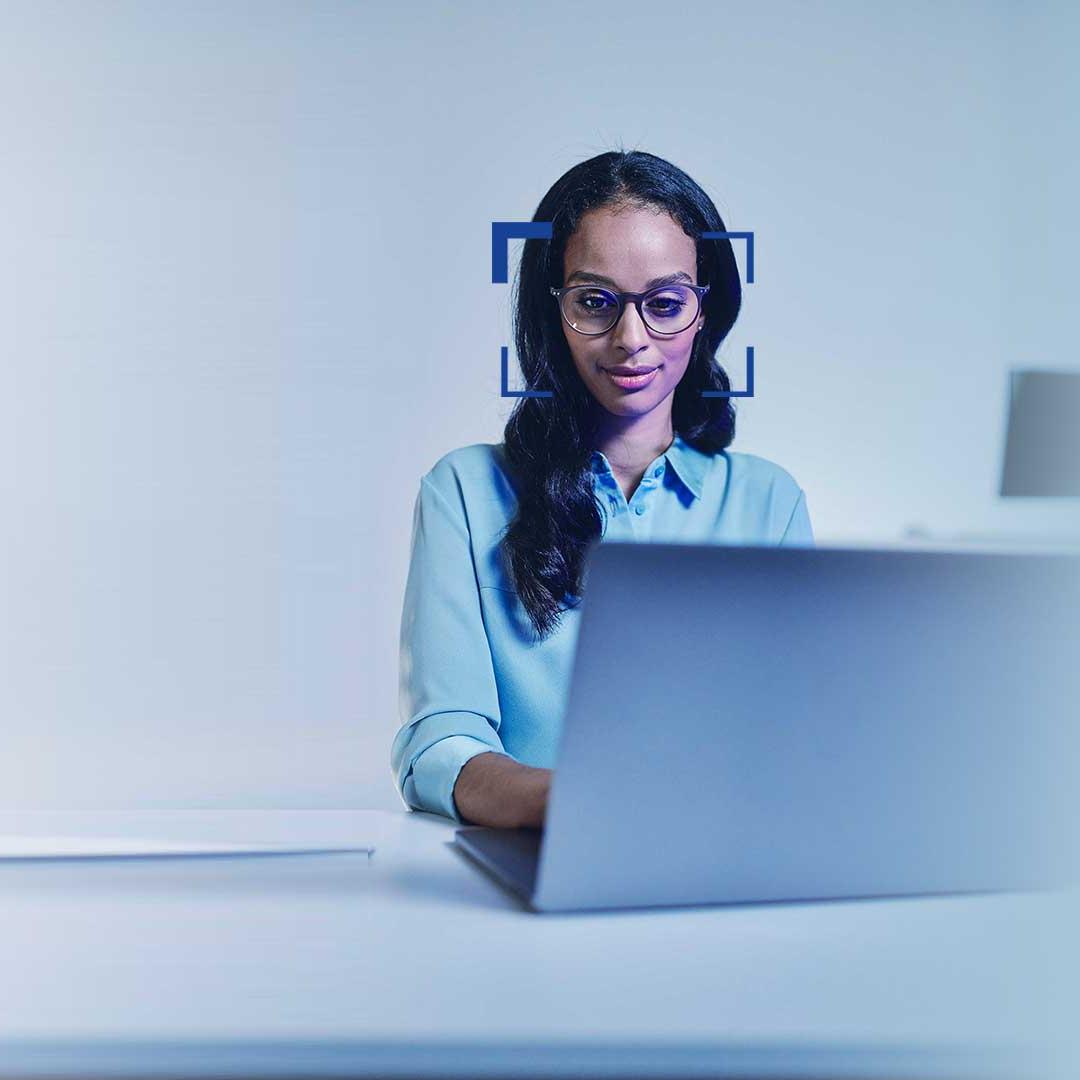 O femeie cu părul negru și ochelari de vedere privește un laptop zâmbind