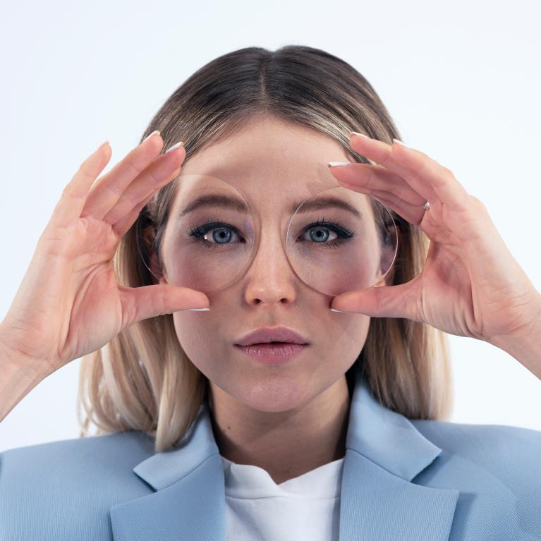 O tânără blondă ține lentilele în fața ochilor pentru a se înfățișa fără niciun efect vizual amuzant, din datorită lentilelor monofocale ZEISS ClearView.