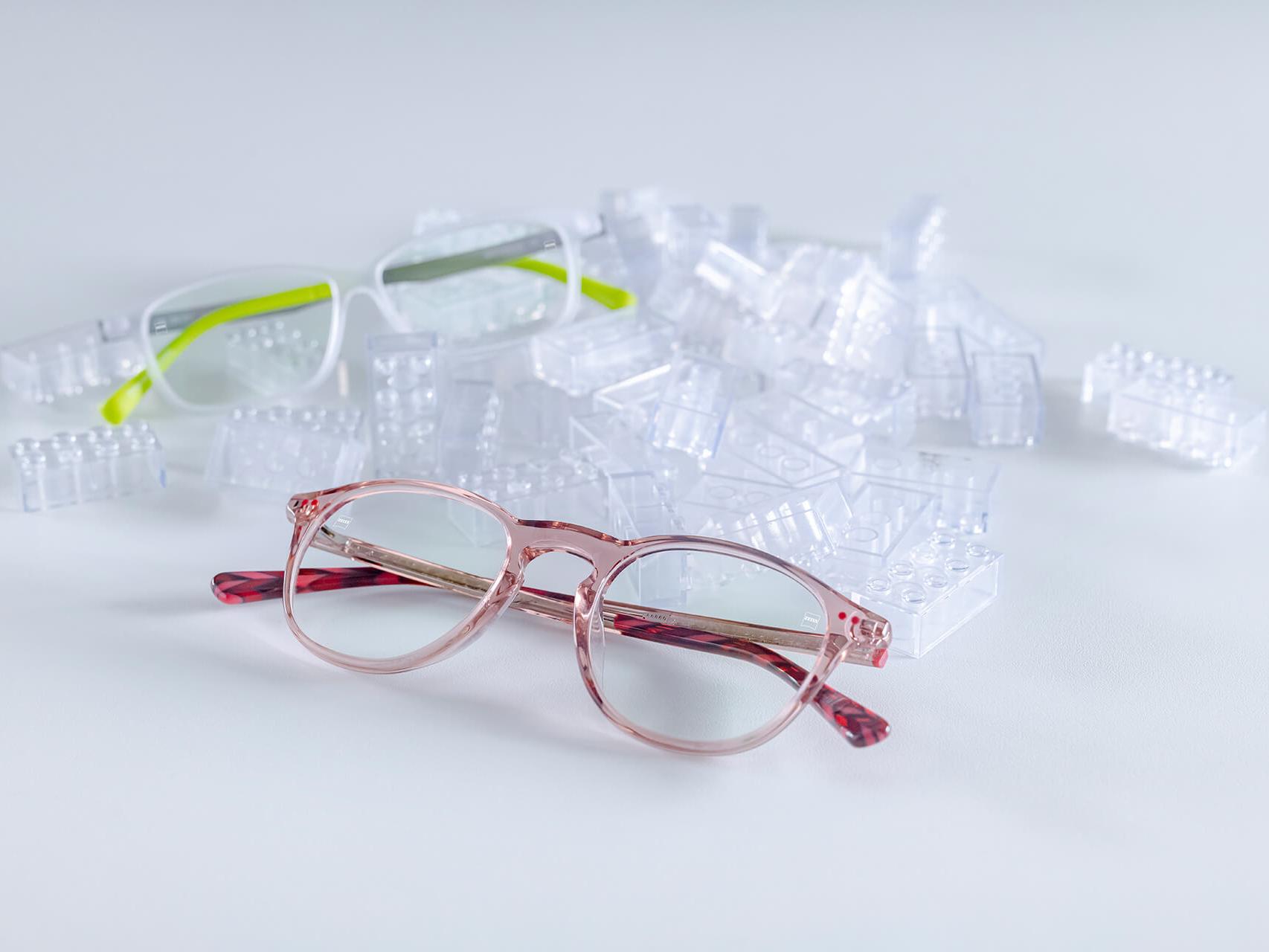 Două perechi de ochelari de ochii cu lentile ZEISS și tratament pentru copii DuraVision® Kids. Ochelarii sunt amplasați între cărămizile transparente ale copiilor.