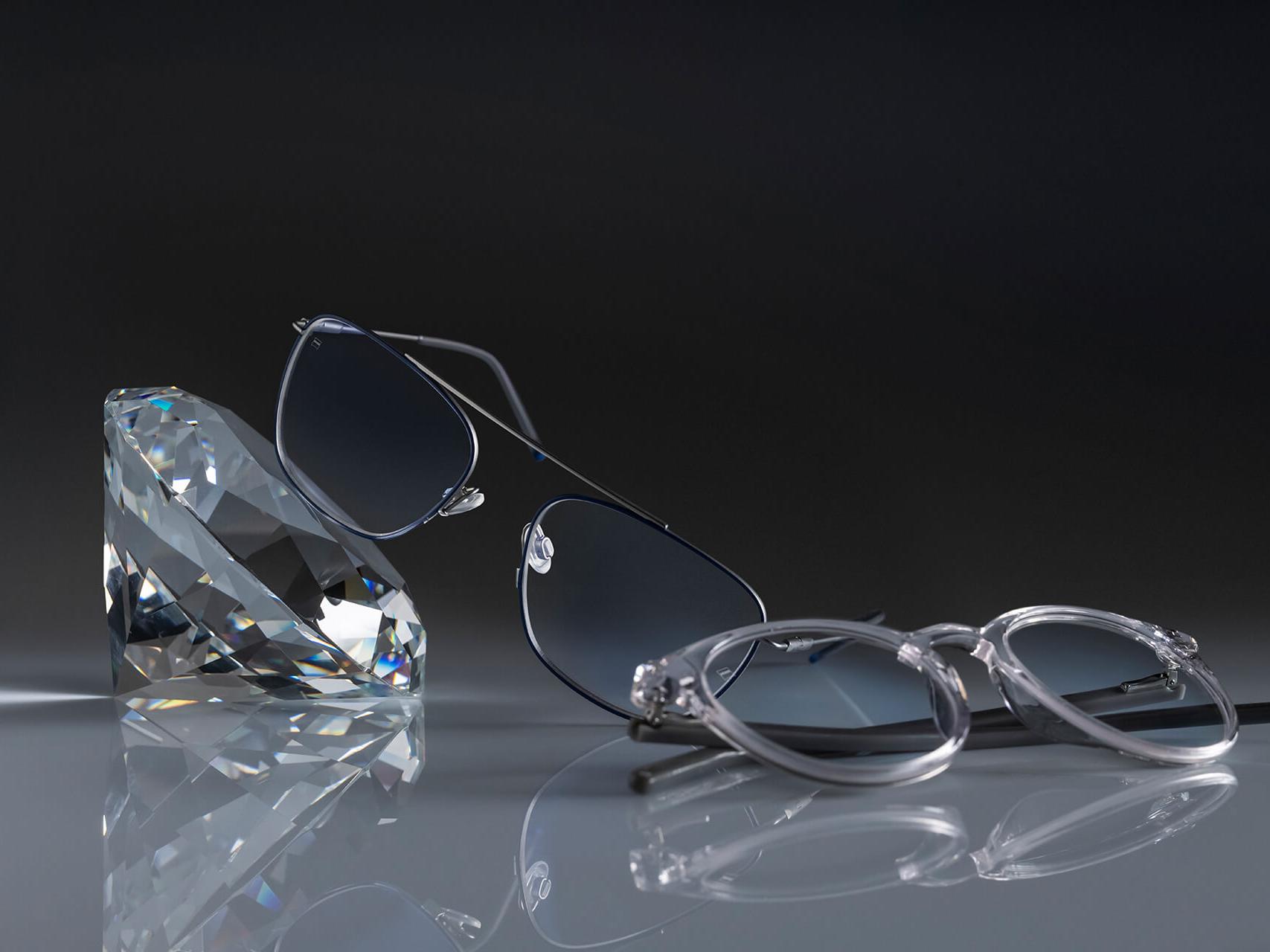 Două perechi de ochelari cu lentile ZEISS care au cel mai dur tratament ZEISS de până acum - DuraVision® Platinum. O pereche de ochelari se sprijină pe un cristal, cealaltă stă plată pe pământ.  Ambele sunt prevăzute cu lentile incolore, fără reflexii albăstrui.