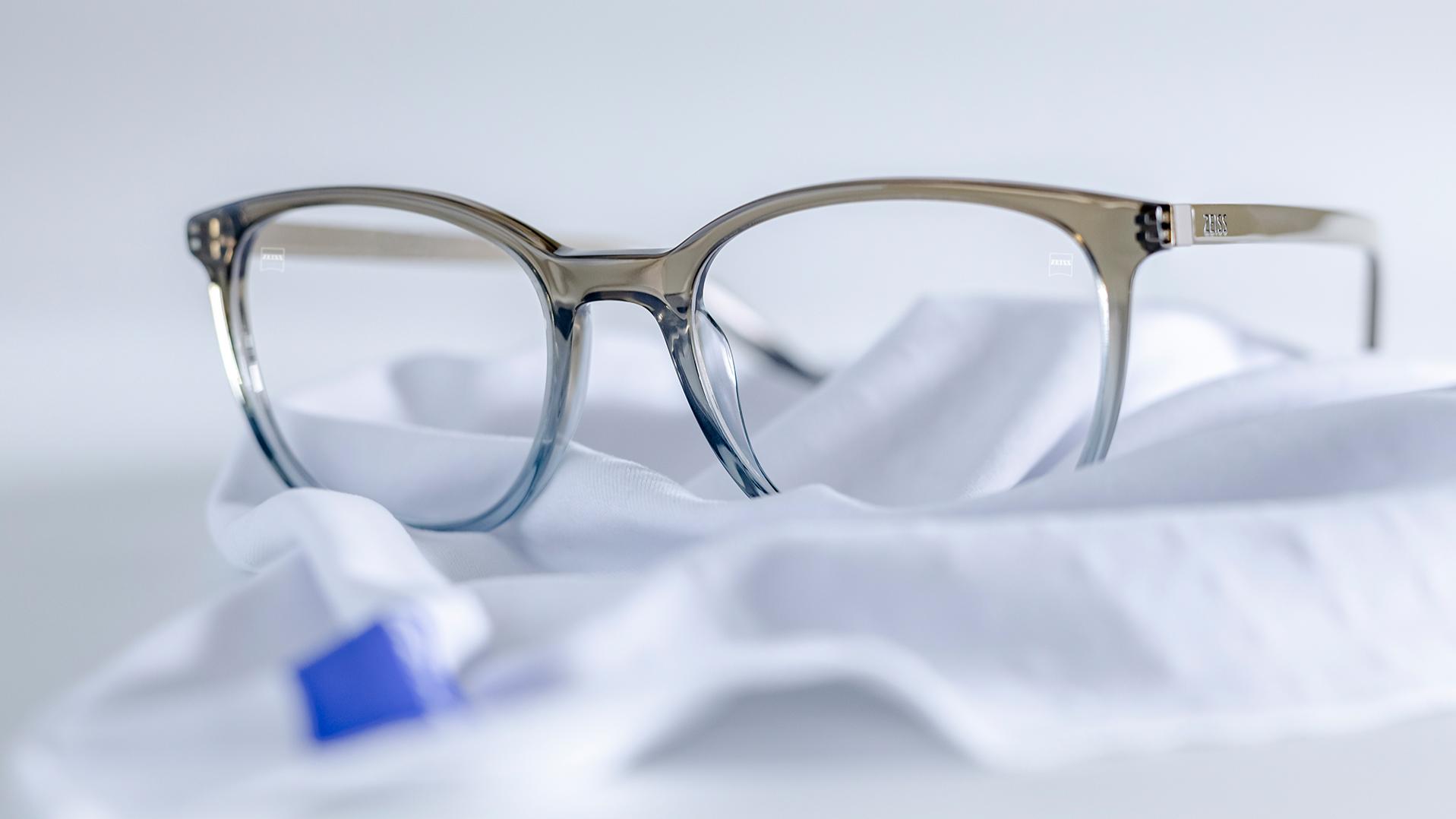 O pereche de ochelari de vedere cu rame gri-albastre și lentile ZEISS cu tratament DuraVision® se află pe o cârpă albă din microfibră.