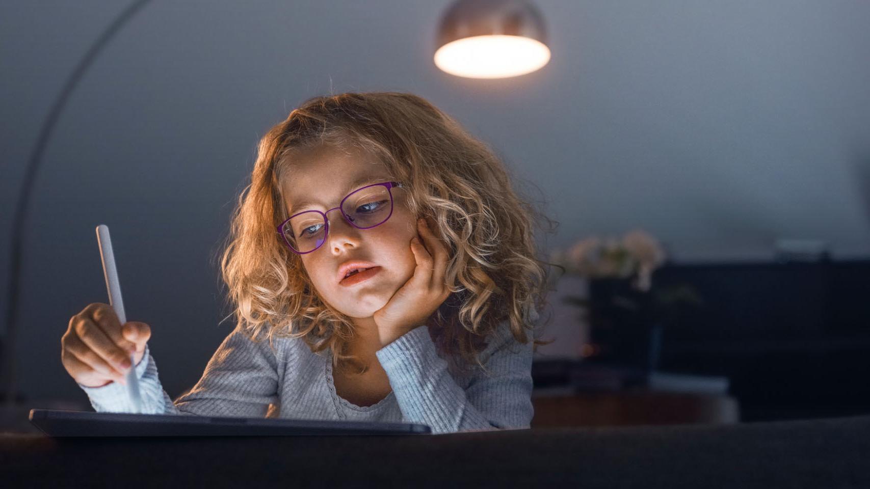 O fetiță într-o cameră de zi întunecată se uită la o tabletă.