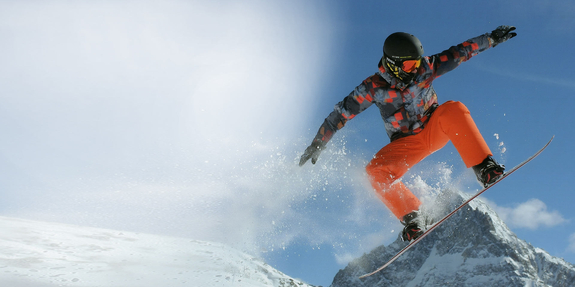 Un bărbat sare cu un snowboard, poartă o cască de schi neagră și ochelari de protecție pentru zăpadă ZEISS cu tehnologie Interschimbabilă pentru protecție