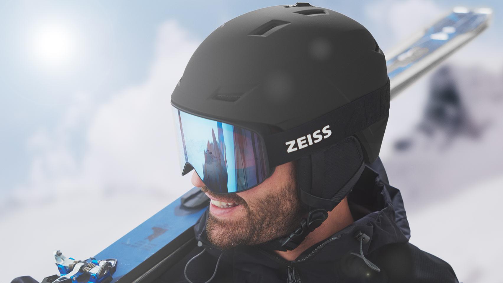 Un bărbat poartă o cască neagră cu ochelari de protecție pentru zăpadă ZEISS tip oglindă albastră, are schiurile pe umărul drept.