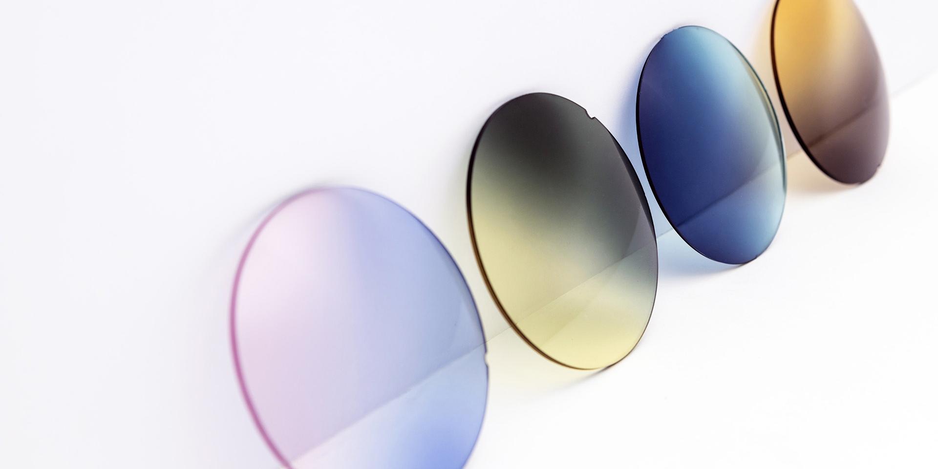 Diferite lentile colorate de ochelari de soare stând pe o suprafață albă: culori degrade roz-violet, galben-gri, albastru și maro.